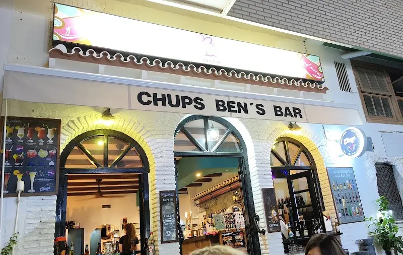 Fachada iluminada de Chups Ben's Bar de noche, con arcos blancos y carta de cócteles