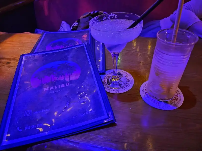 Cartas de Malibu bajo luz azul junto a un margarita y un combinado en vaso alto