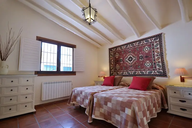 Dormitorio con dos camas, techo de vigas blancas, alfombra colgada y cómoda