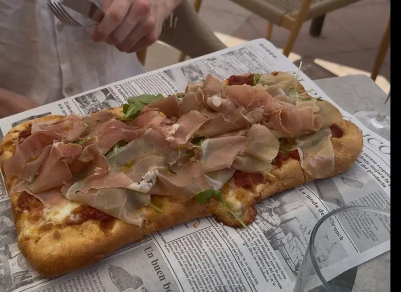 Pizza rectangular con jamón curado, rúcula y tomate sobre papel de periódico