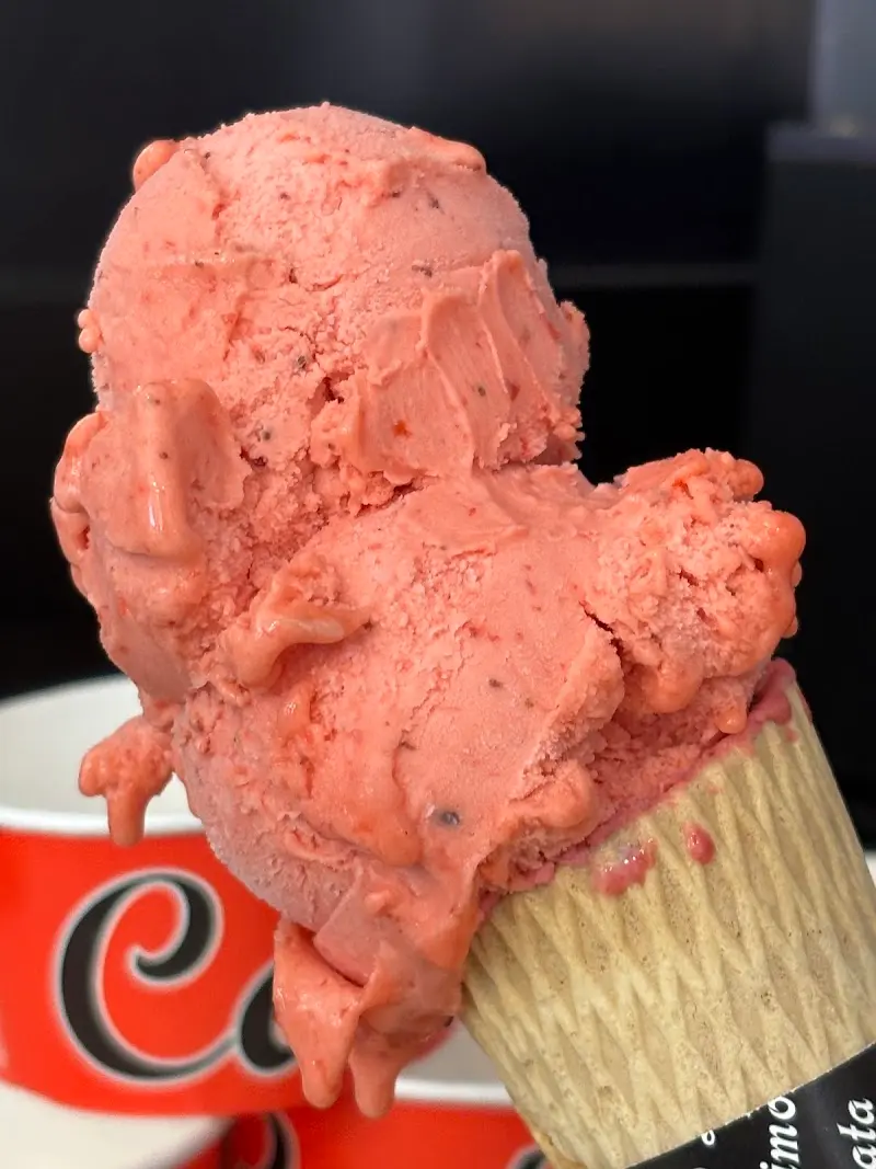 Cucurucho con bolas de helado de fresa ante una tarrina con el logotipo de la casa