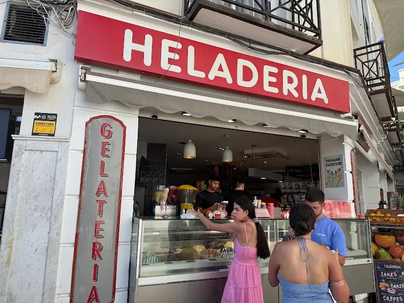 Clientes eligiendo helado en la vitrina, con columna de Gelateria y carteles de crepes