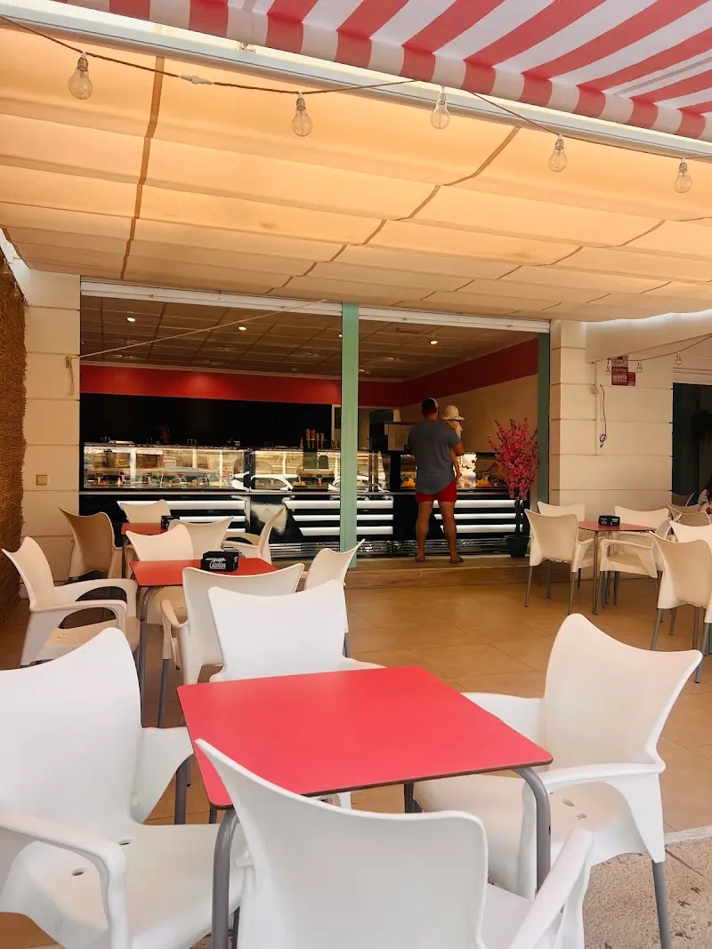 Terraza bajo el toldo con mesas rojas, sillas blancas y el mostrador de helados al fondo