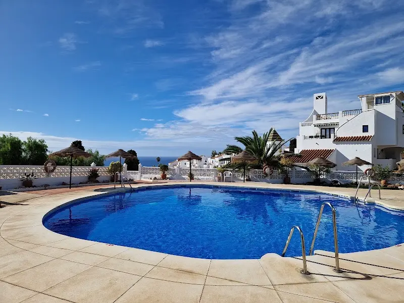 Piscina curva con sombrillas de paja, casas blancas y el mar a lo lejos