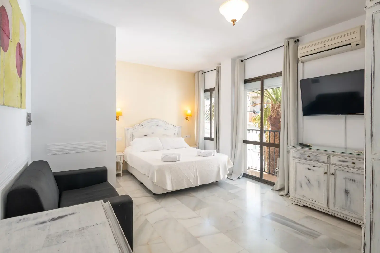 Habitación con cama de matrimonio, sofá, televisor en la pared y ventanal al balcón