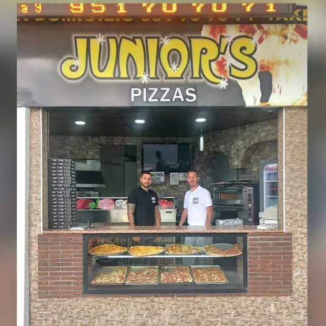 Mostrador de Junior's Pizzas con pizzas en porciones en la vitrina y dos pizzeros