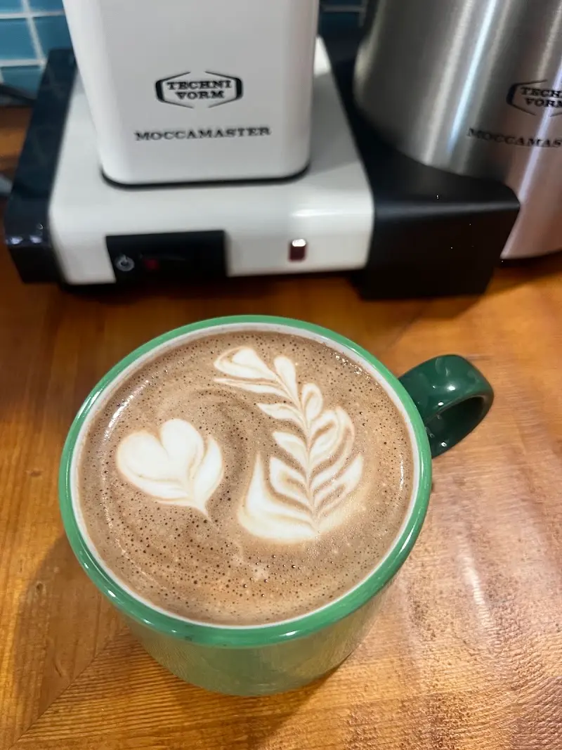 Café con leche con arte latte en taza verde junto a una cafetera Moccamaster