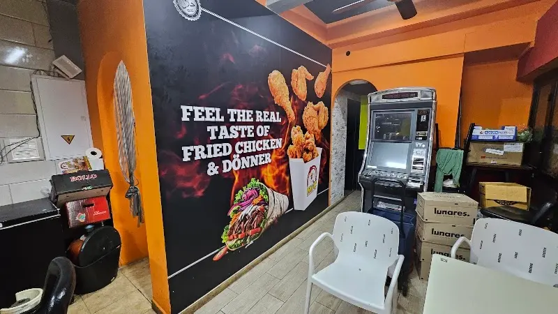 Interior con mural de pollo frito y döner, mesas blancas y una máquina recreativa
