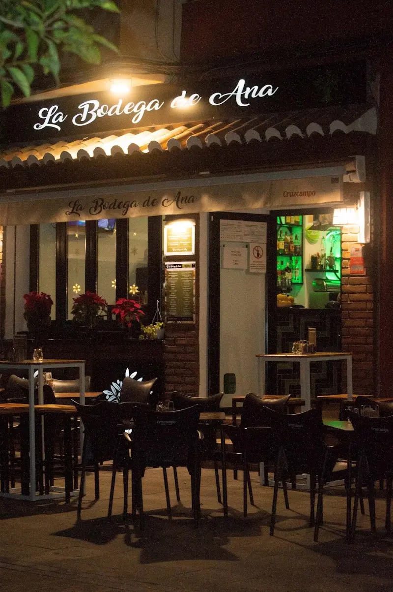 Fachada iluminada de noche con el rótulo del bar y la terraza de mesas vacías