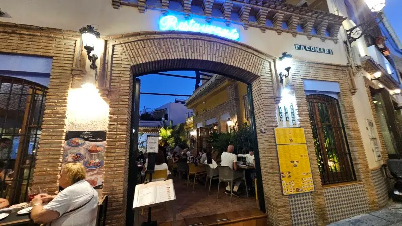 Entrada en arco de ladrillo con rótulo de neón y patio con clientes al anochecer