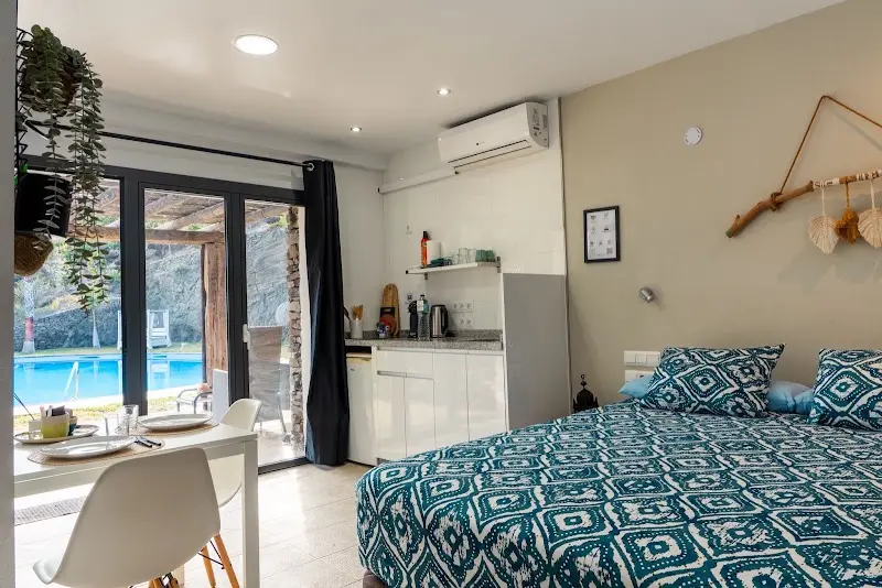 Estudio con cama de matrimonio, cocina pequeña, mesa blanca y puerta de cristal a la piscina