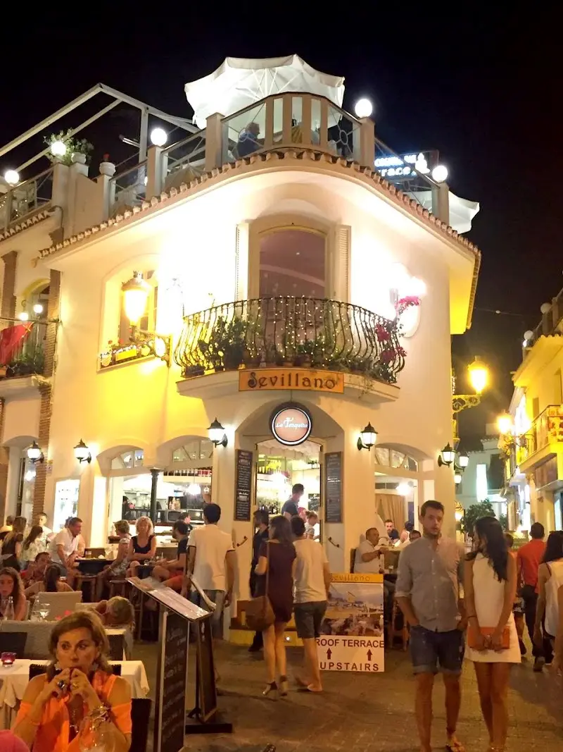Fachada iluminada de noche con balcón de forja, terraza llena y cartel de azotea arriba