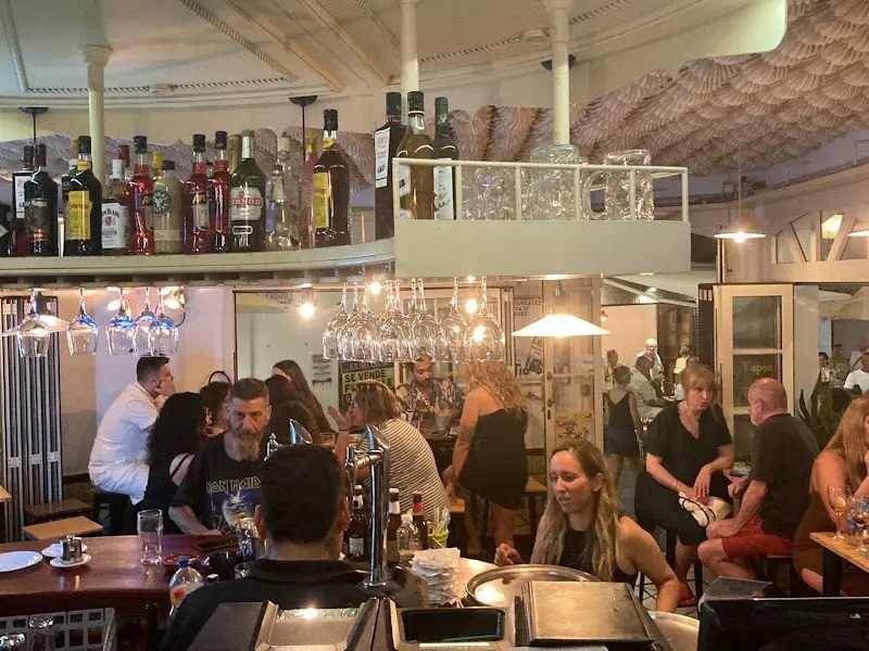 Clientes en la barra y las mesas altas por la noche, bajo un botellero con copas colgadas