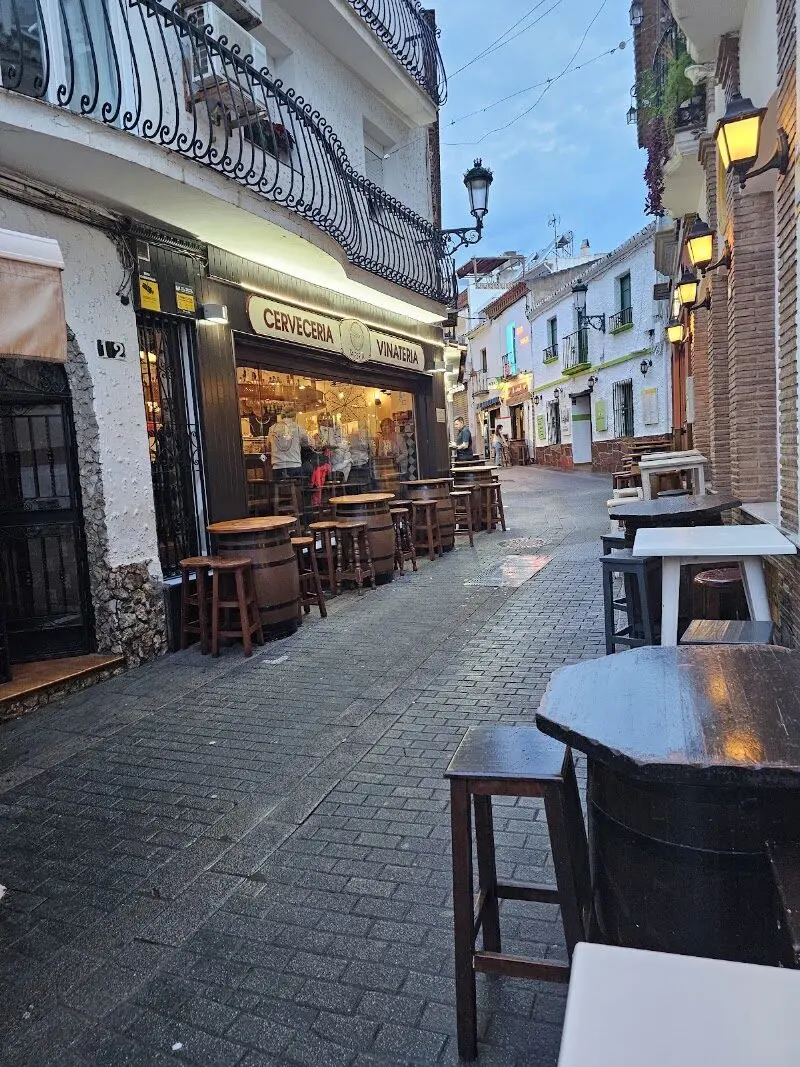 Cervecería vinatería en una calle empedrada al anochecer, con mesas de barril y taburetes en la acera