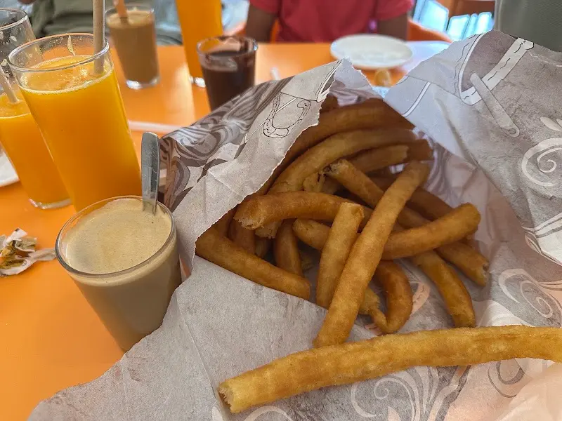 Churros en su papel con café con leche y zumos de naranja en una mesa naranja