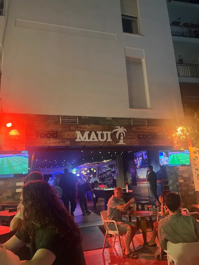 Fachada del Maui Cocktail & Food Bar de noche, con terraza, pantallas de fútbol y clientes