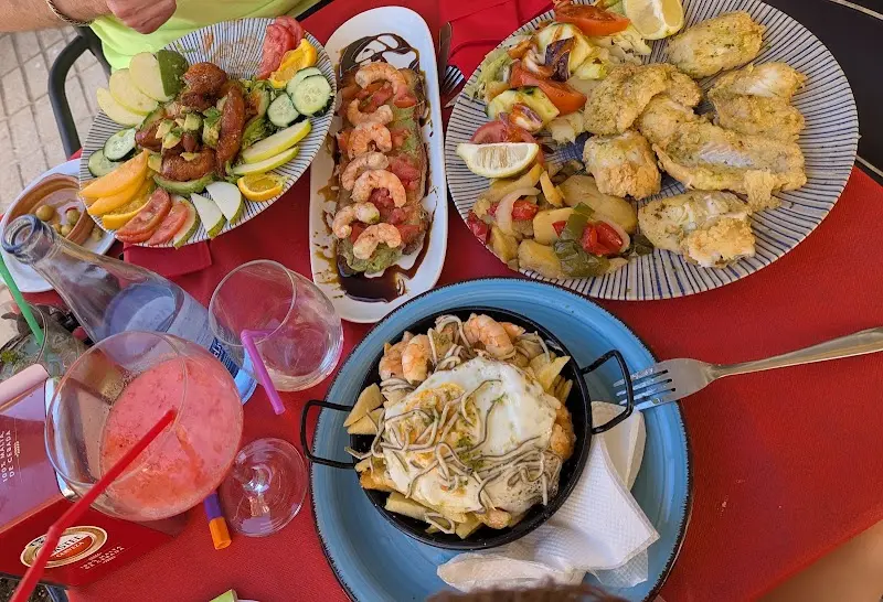 Mesa con varios platos: pescado empanado, gambas, ensalada y huevos rotos con gulas