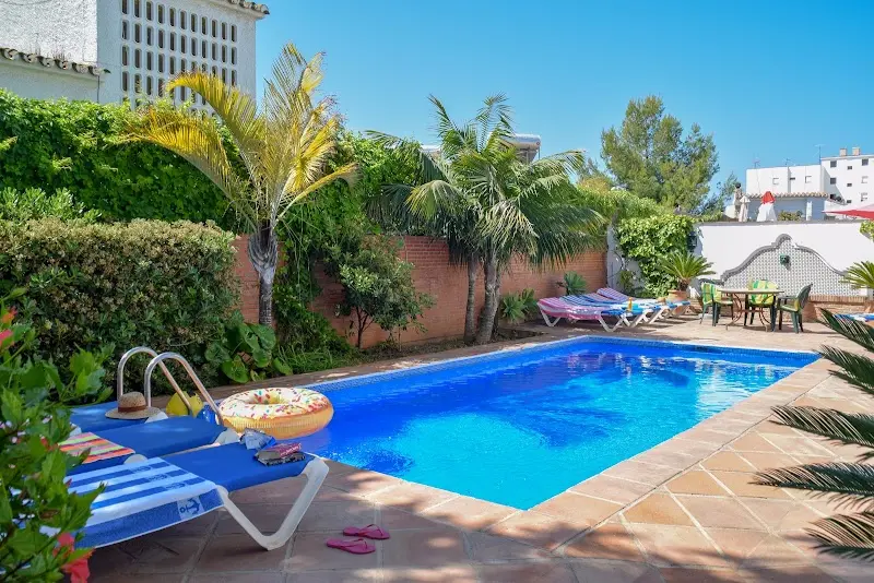 Piscina rodeada de palmeras y setos, con tumbonas, flotador y mesa de jardín