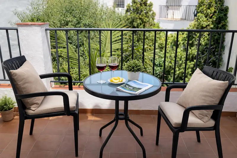 Balcón con mesa de cristal, dos sillones con cojines y copas de vino frente a la vegetación