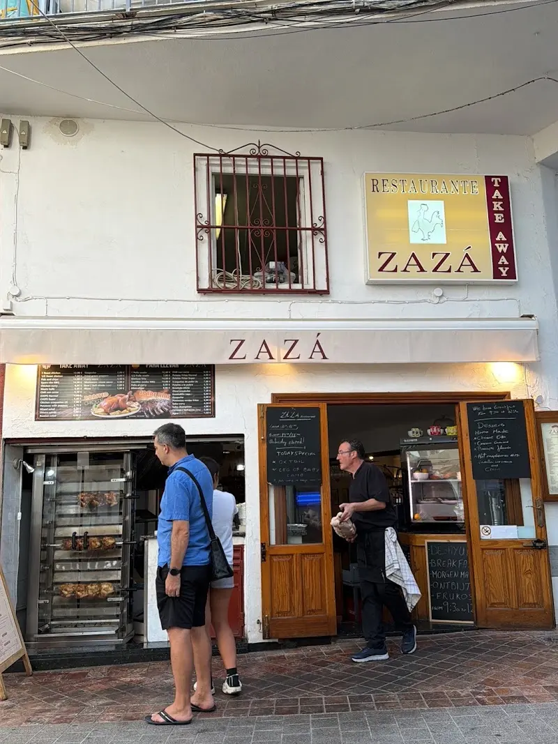 Fachada de New Pollo Asado Zazá con el asador de pollos a la vista y pizarras en la puerta
