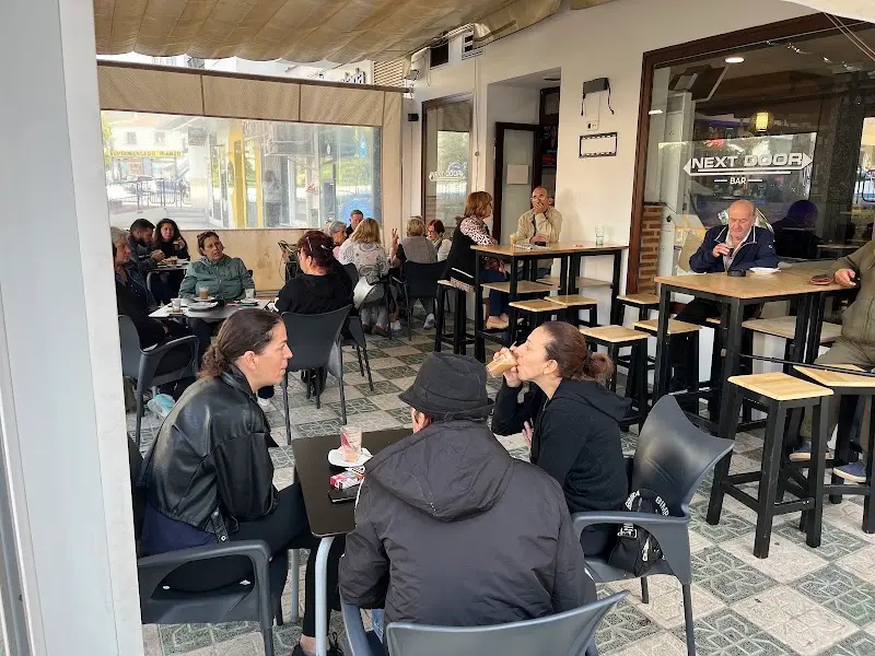 Terraza cerrada del bar con clientes tomando café en mesas altas y bajas