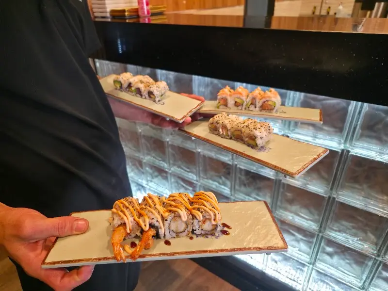 Camarero sosteniendo cuatro bandejas de uramakis, uno con langostino en tempura y salsa