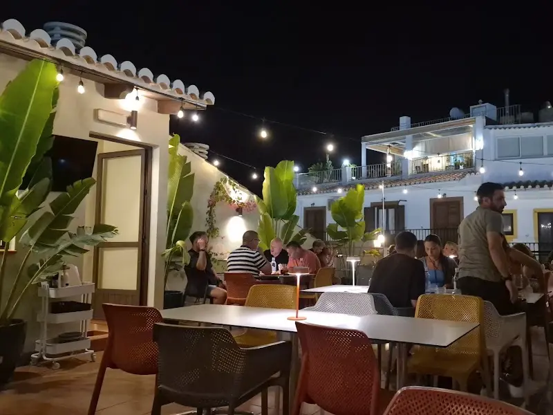 Terraza en la azotea de noche con plantas, guirnaldas de luces y comensales en las mesas