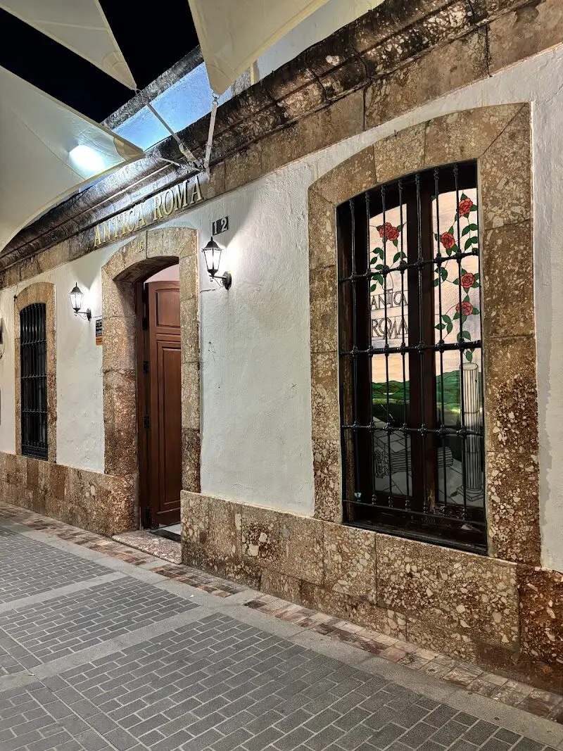 Fachada encalada con marcos de piedra, puerta de madera y vidriera con el nombre Antica Roma