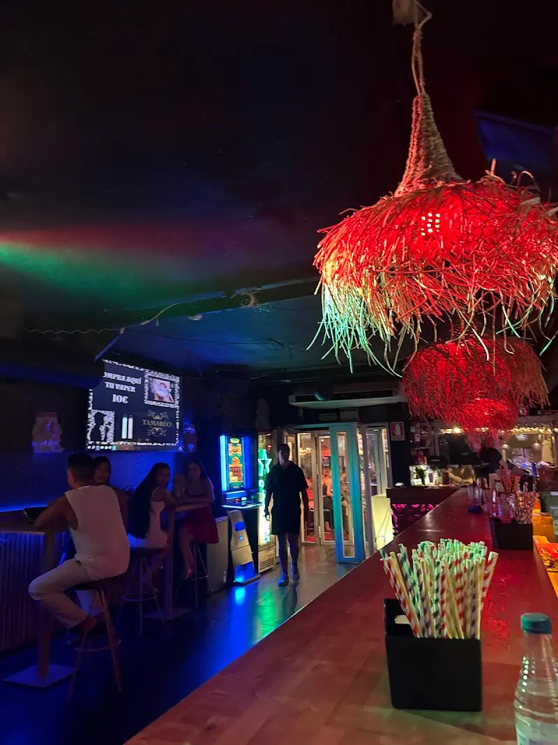 Interior de noche con lámparas de rafia rojas, clientes en mesas altas y pantalla de karaoke