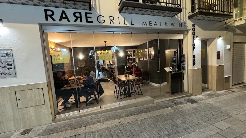 Fachada acristalada de Rare Grill Meat & Wine de noche, con comensales en el interior