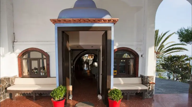 Entrada encalada del restaurante con puerta de madera, ventanas de arco y macetas rojas