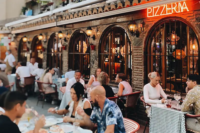 Terraza de la pizzería con clientes cenando bajo el rótulo luminoso y ventanales en arco