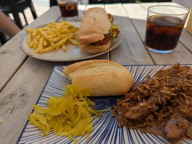 Carne mechada con pan y encurtidos, y un bocadillo con patatas fritas en una mesa de madera
