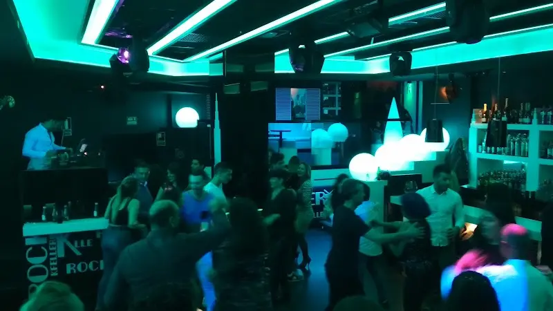 Pista llena de gente bailando bajo luces turquesa, con el DJ pinchando a la izquierda