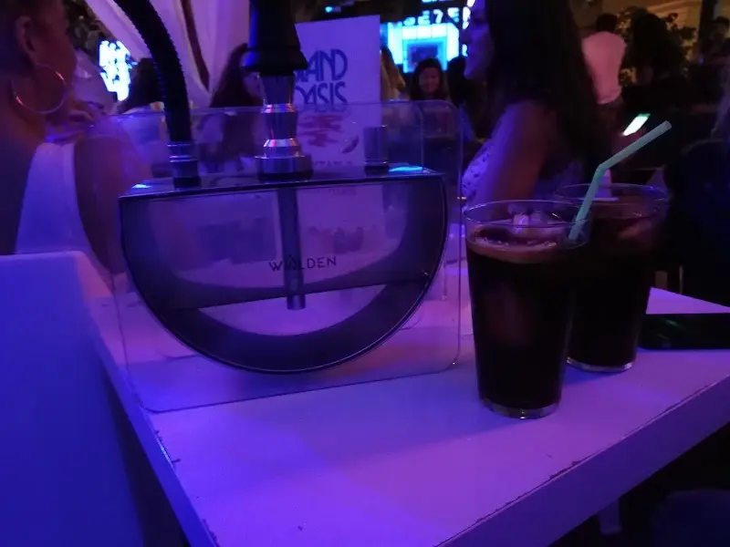 Cachimba transparente y dos refrescos sobre una mesa blanca con luz morada