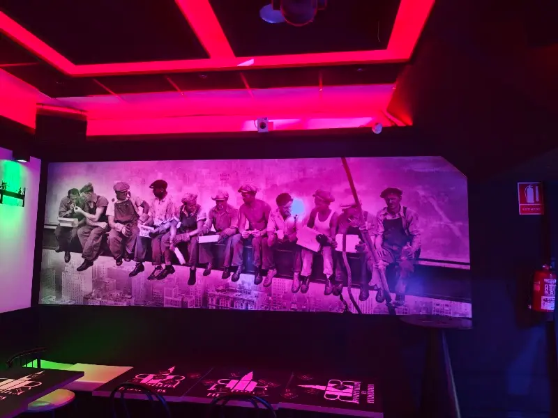 Mural de obreros sentados en una viga sobre la ciudad, bajo luces de neón rosas
