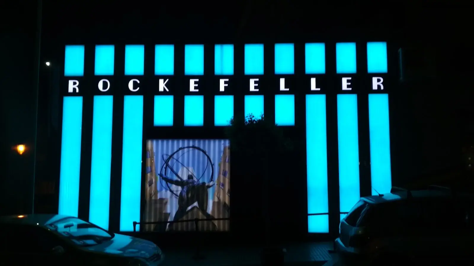 Fachada de Rockefeller de noche, con su nombre sobre columnas iluminadas en turquesa
