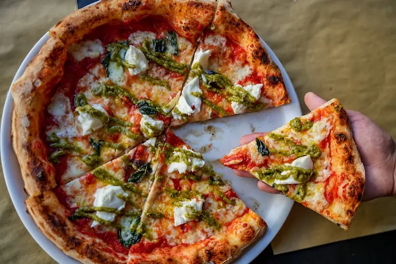 Pizza con tomate, mozzarella fresca y pesto, con una porción en la mano