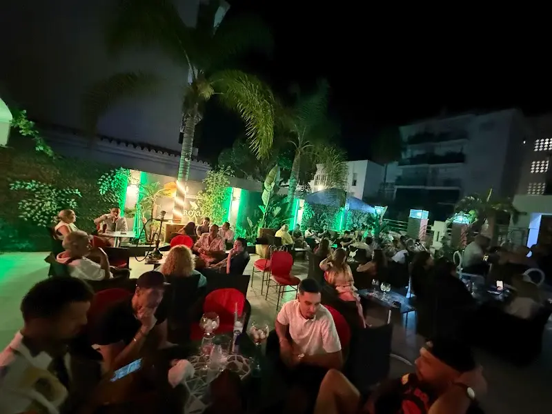 Terraza de Savage llena de gente de noche, con palmeras y luces verdes en la Plaza Tutti Frutti