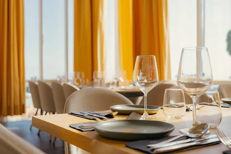 Mesa puesta con platos grises, copas de vino y cubiertos junto a las cortinas amarillas