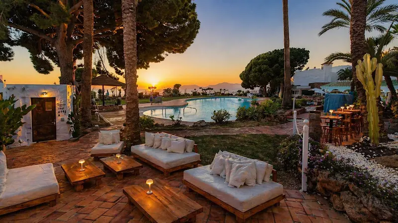 Jardín con sofás blancos y velas junto a la piscina, con el sol poniéndose tras las palmeras