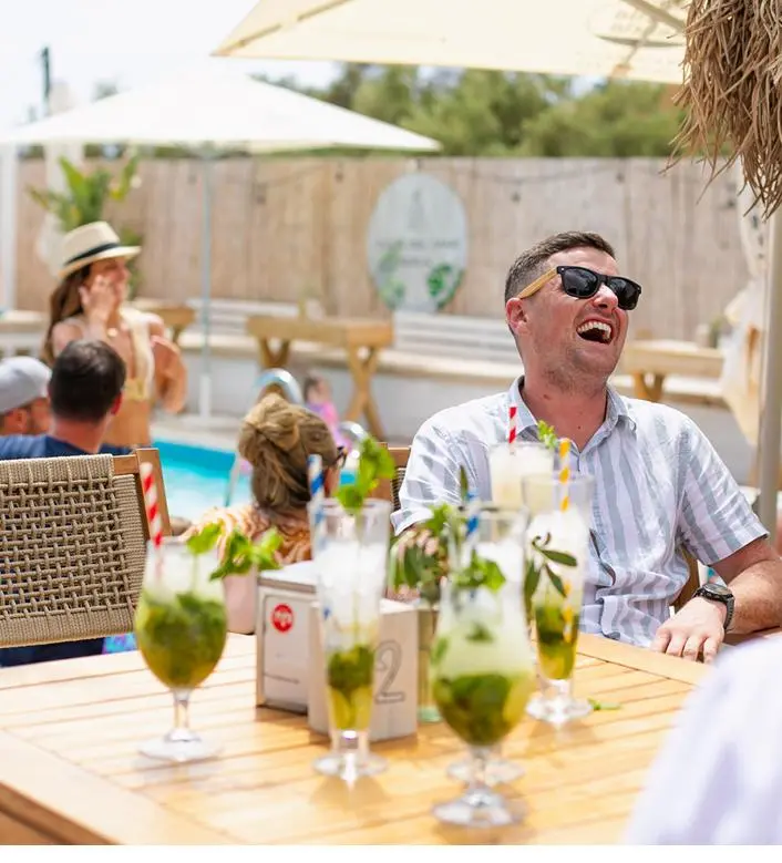 Clientes riendo en una mesa con mojitos junto a la piscina del pool club