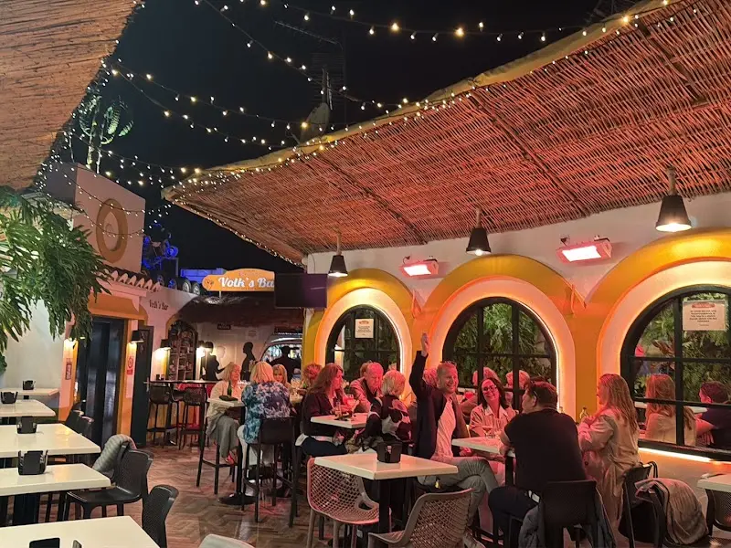 Terraza de noche llena de clientes bajo el cañizo, con arcos amarillos iluminados