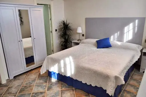 Dormitorio con cama de matrimonio, cojín azul, armario blanco con espejo y suelo de baldosas