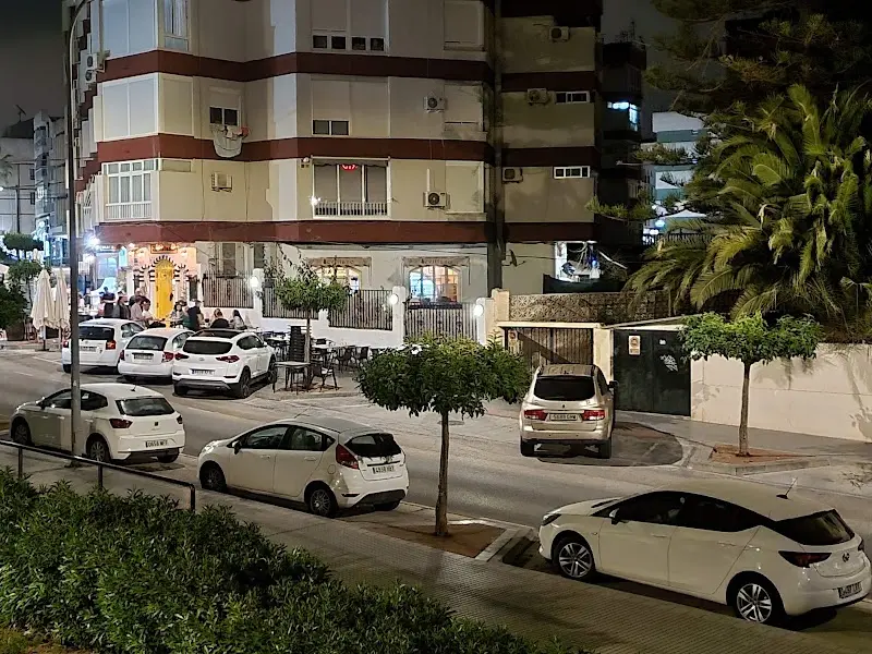 Calle de noche frente al edificio, con coches aparcados y gente en la terraza de un bar