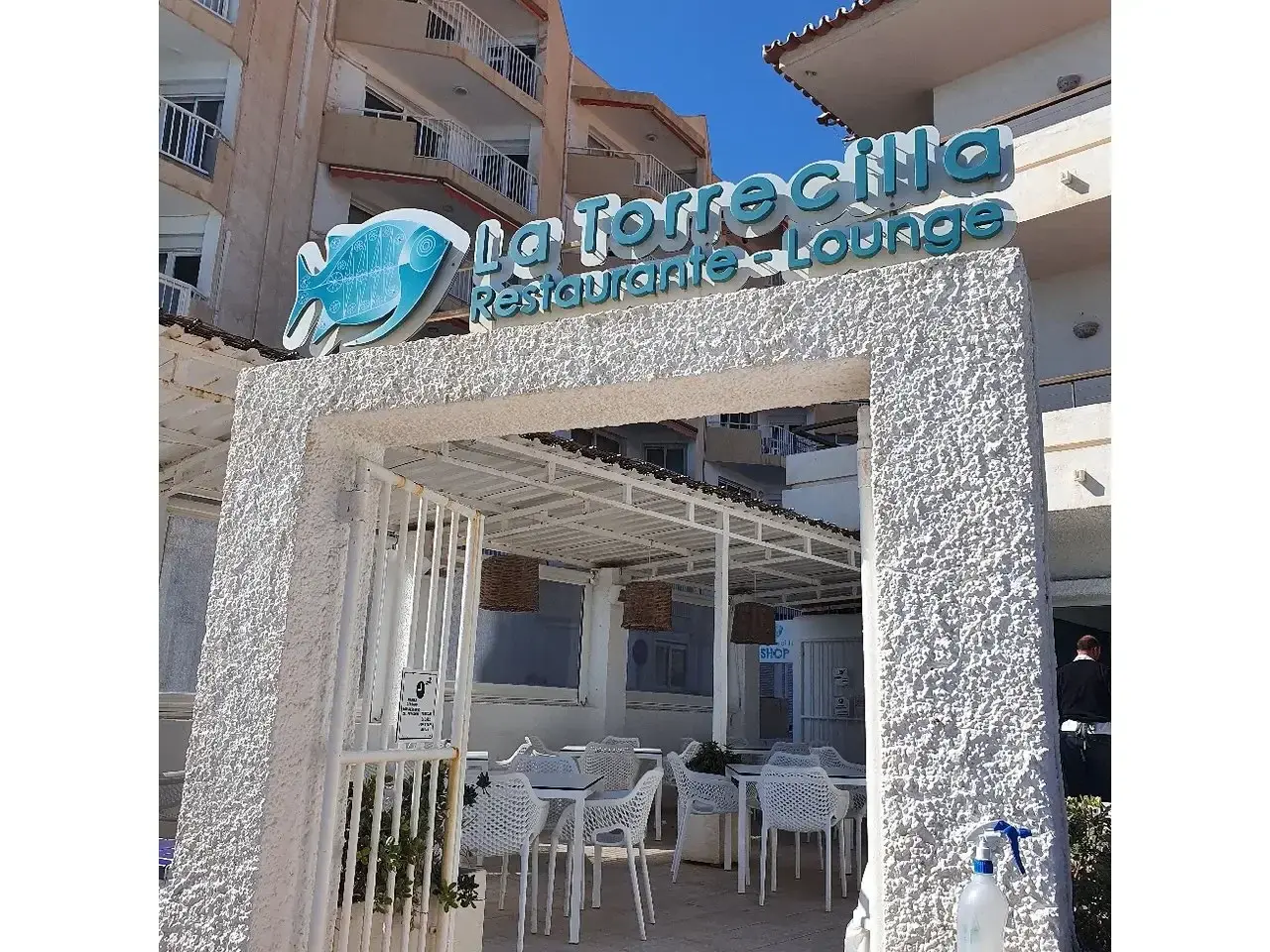 Entrada del Restaurante La Torrecilla con su rótulo de pez azul «Restaurante - Lounge» y la terraza al fondo